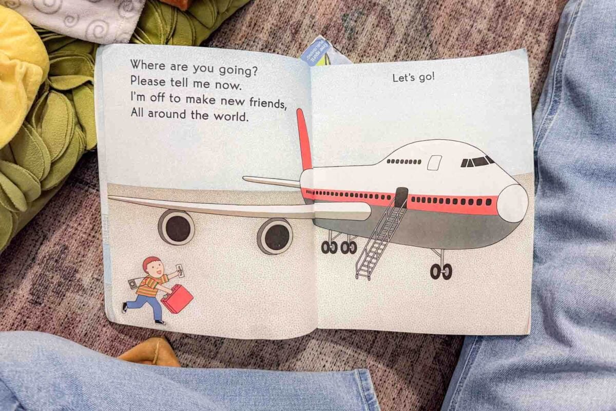 Open children’s board book shows a large jumbo double-decker airplane with boarding stairs while a small child runs toward it holding a suitcase, illustrating excitement for a long haul flight with a toddler. The page text reads “Where are you going. Please tell me now. I’m off to make new friends, All around the world.” and “Let’s go.”