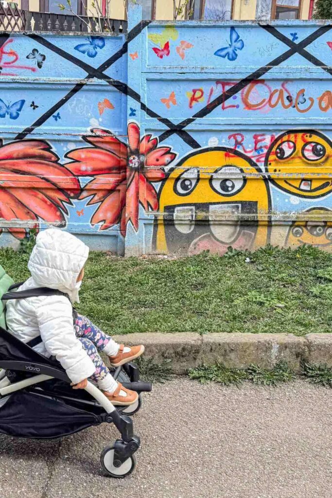 Toddler in white puffy coat seated in a Babyzen Yoyo stroller looking at colorful street art in Brasov, Romania, showing urban sightseeing with a compact travel stroller.