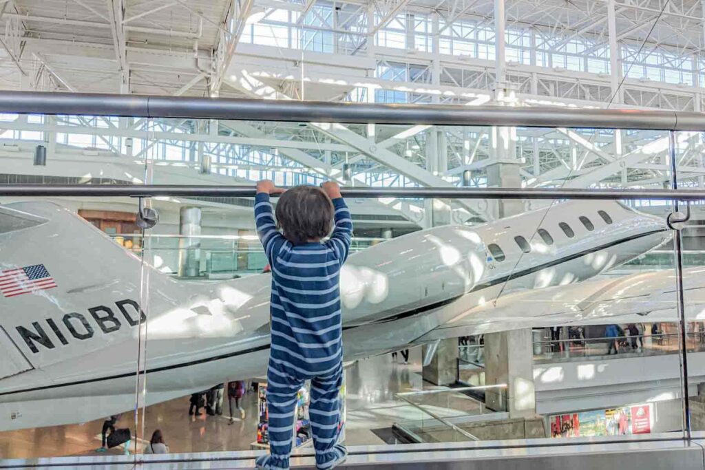Toddler standing at the Denver airport mezzanine railing checking out the hanging real-life jet plane while waiting for a flight. Exploring the airport can help with burn off toddler energy before flying.
