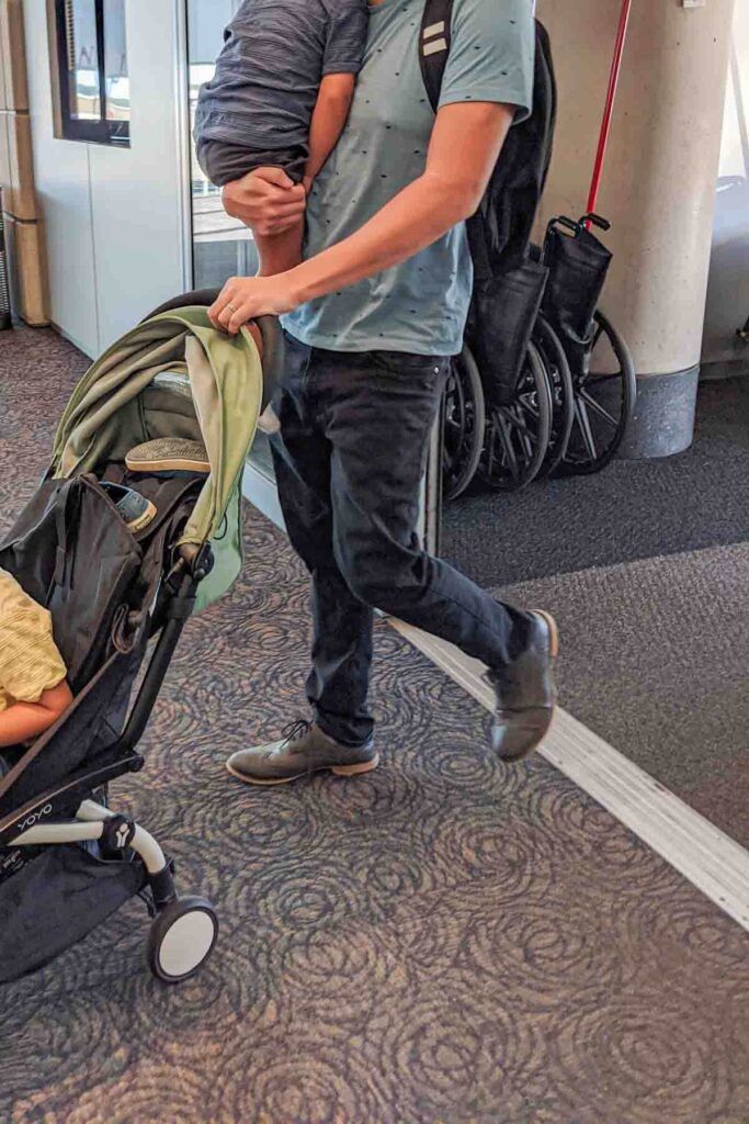 Dad holding a toddler over his shoulder with one arm while pushing a Babyzen Yoyo stroller with the other hand, demonstrating maneuverability and ease of use showing why it’s one of the best travel strollers.