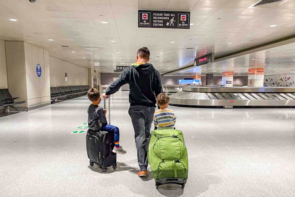 A dad walks through an empty airport baggage claim pulling a small black rolling suitcase and a large bright green rolling suitcase with young children sitting on top of each one. The image captures a real moment of family travel and managing luggage efficiently with kids.
