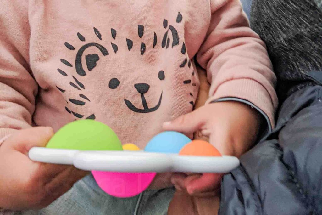 Toddler wearing a pale salmon sweatshirt with a modern hedgehog pattern on it holding a colorful Dimpl pop toy during a flight. Simple toys can easily entertain a toddler on a long flight.