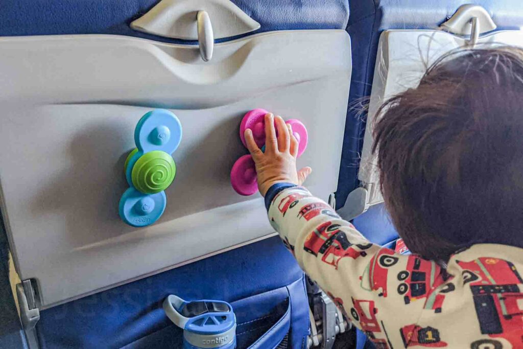 Toddler wearing firetruck pjs pressing colorful Fat Brain Toys Whirly Squigz suction toys onto a stowed airplane tray table during a long haul flight with a toddler, showing a fun toy to entertain a toddler on a plane.