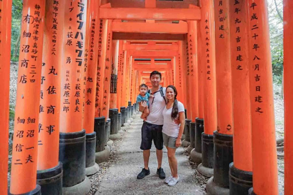 Family standing beneath bright red orange torii gates at the famous Fushimi Inari shrine in Kyoto, Japan while holding their baby. Iconic cultural stop when traveling to Japan.