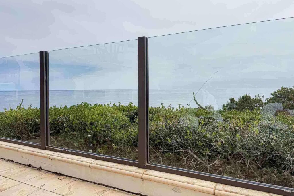 View of the ocean off Rancho Palos Verdes, California through a glass barrier fence at Terranea Resort, bordered by coastal shrubs and overcast skies.