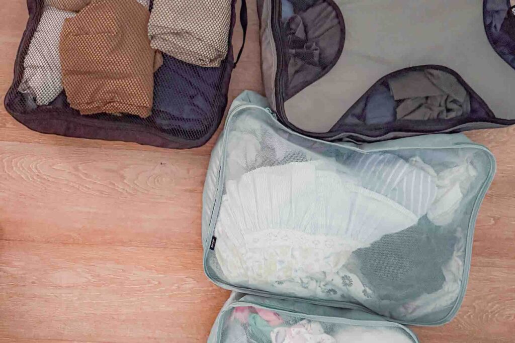 Gray and aqua mesh packing cubes filled with folded clothing are laid out on a wooden floor in preparation for a family vacation. 