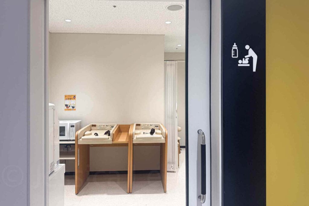 Entrance to a baby care room in Haneda airport in Tokyo, Japan with a baby bottle icon and adult changing a baby icon with a view inside the room of changing tables, a microwave and feeding area visible inside. Practical amenity that makes traveling to Japan with a baby easier.