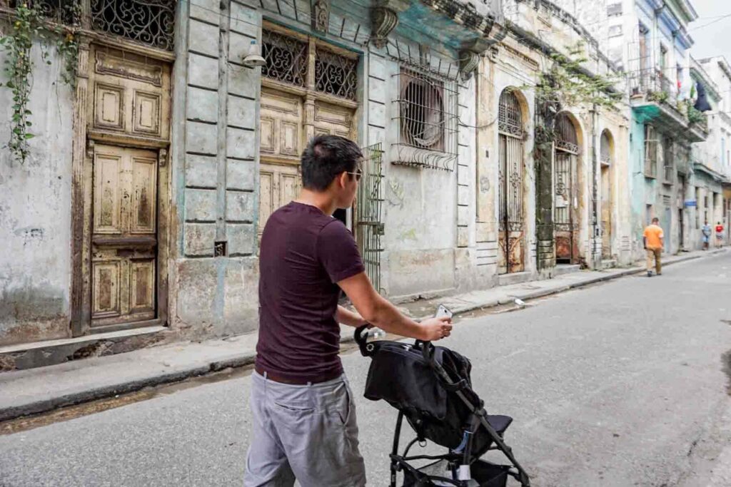 Dad wearing a burgundy tshirt and light gray shorts pushing a black stroller down a street of neoclassical buildings in Havana, Cuba.
