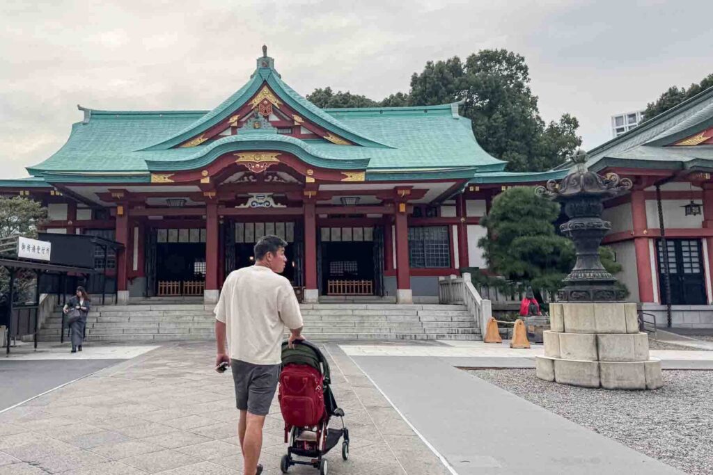 Father pushes a travel stroller with a red backpack hanging from it through the Hie Shrine complex with the main red building with a green roof and gold accepts in Tokyo.