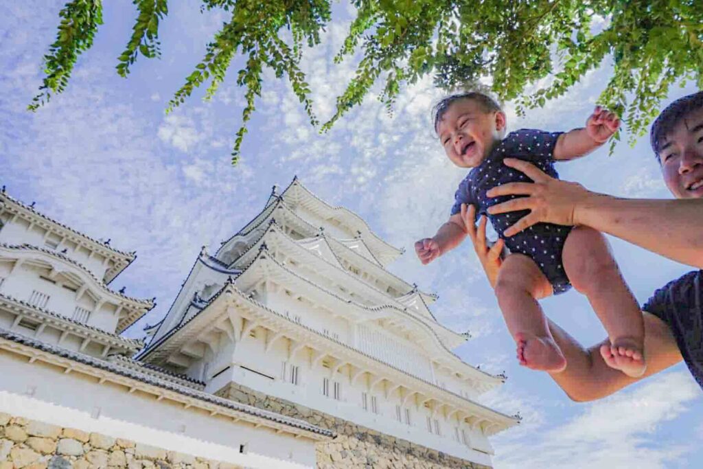 Parent lifting a smiling baby in front of the grand white Himeji Castle under a blue sky. Iconic landmark to visit when traveling to Japan with a baby.
