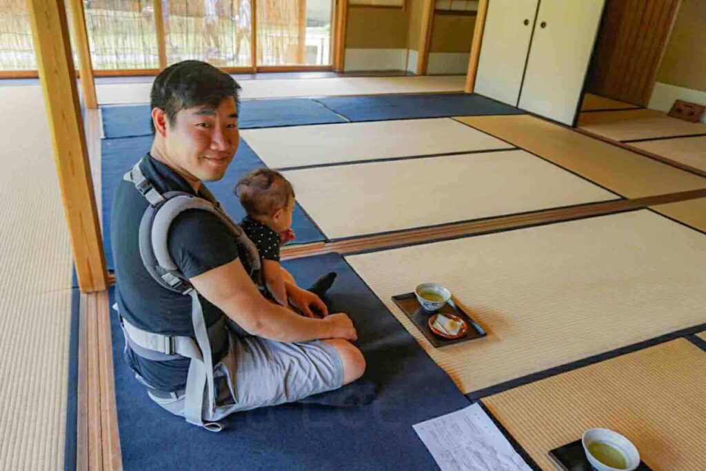 Father sitting on tatami mats with baby in a traditional Japanese room with tea set on a tray in Hemji japanese garden. 