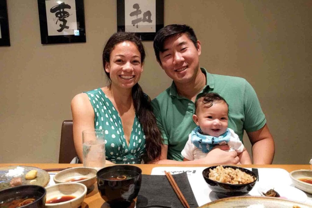Smiling parents sitting at a restaurant table in Kobe, Japan holding their baby with traditional dishes in front of them. Dining out together while traveling to Japan with a baby.