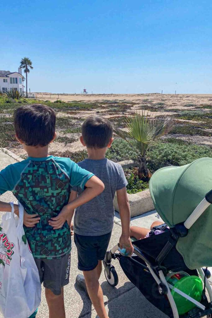 Two kids standing next to a green Babyzen Yoyo travel stroller in Newport Beach, California, showing how a compact stroller fits family outings with siblings.