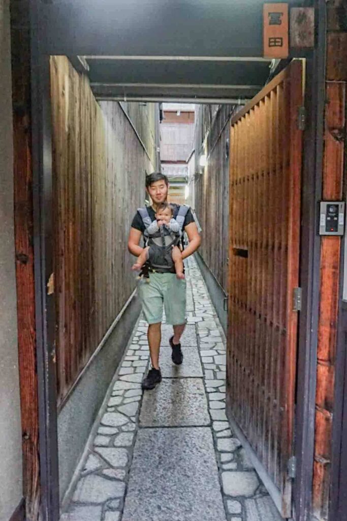 Father walking through a narrow wooden alley in Kyoto while carrying baby on his front in a baby carrier.