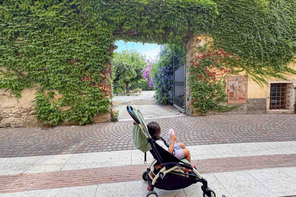 Toddler sitting in a Babyzen Yoyo stroller near a vine covered wall in Lazise, Italy on Lake Garda, showing easier travel days with a lightweight stroller.
