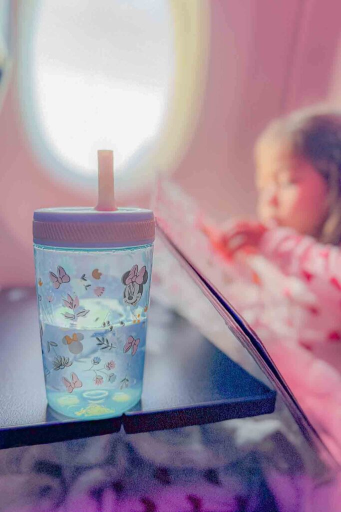 Minnie Mouse pastel blue and pink spill proof sippy cup on an airplane tray table with a toddler blurred in the background on a flight.