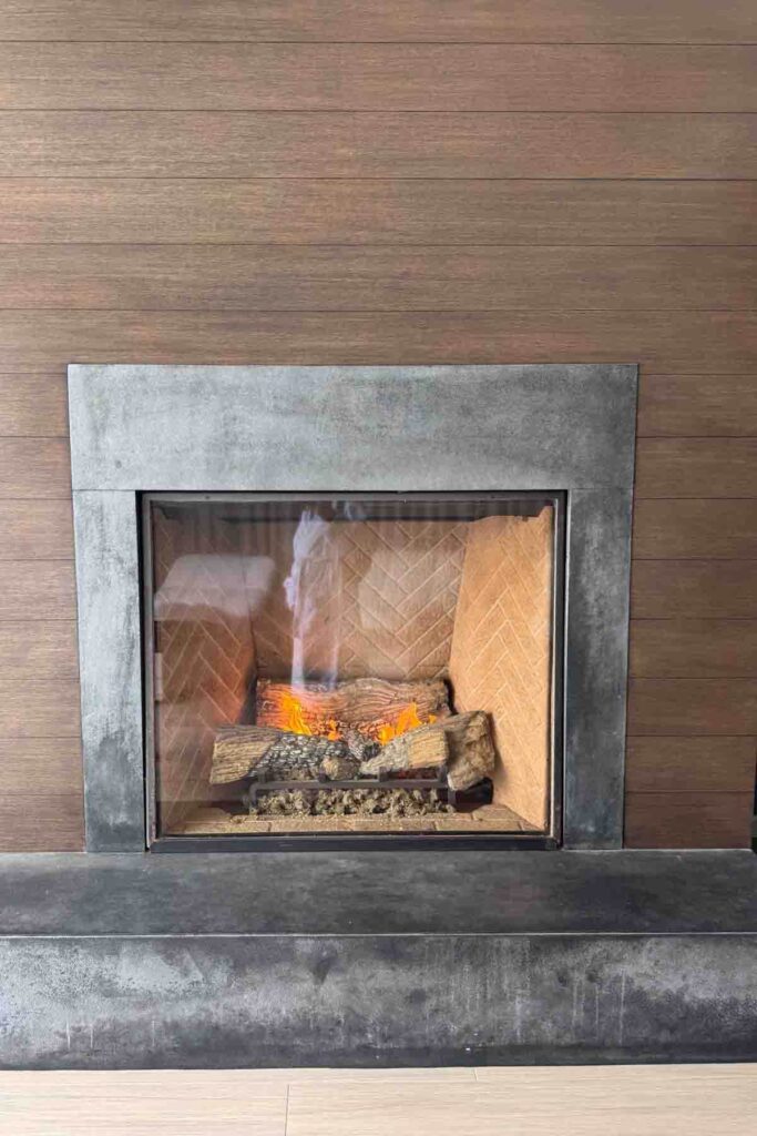 Cozy indoor fireplace framed by modern stone and wood with a slight reflection of a person in a white robe at Terranea Resort. A relaxing space for winding down during a romantic weekend escape.