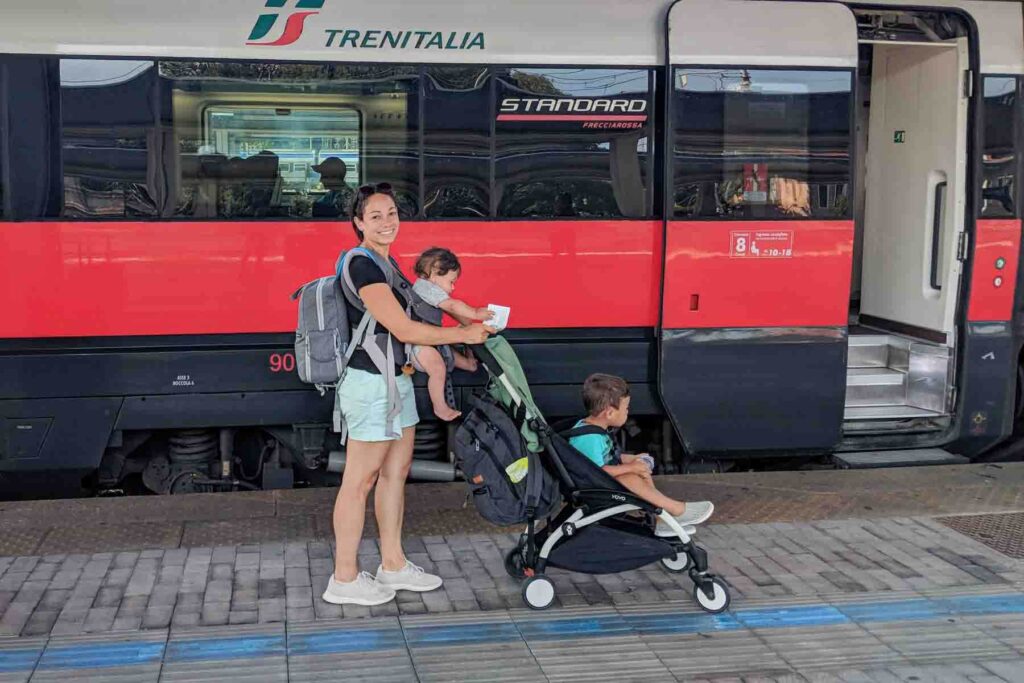 Mom babywearing an infant wearing a backpack while pushing a toddler in a Babyzen Yoyo stroller at an Italian train station in Venice in front of a Trenitalia red train, showing hands on travel with multiple young kids.