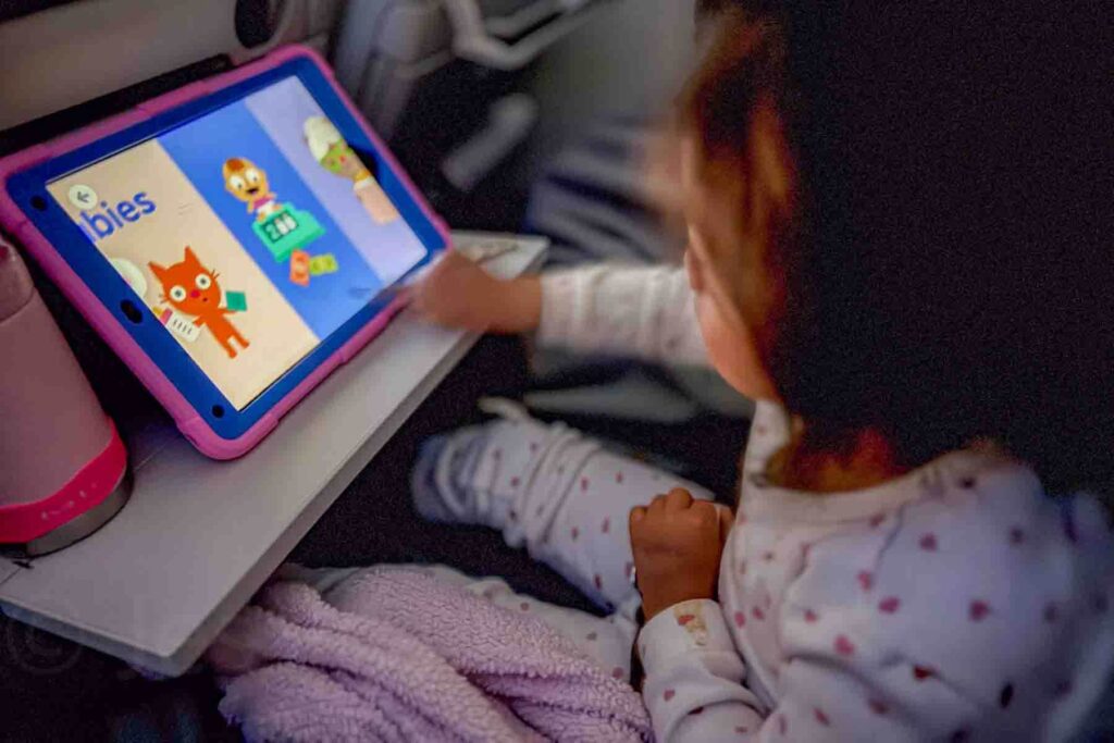 Toddler wearing white pajamas with red and pink hearts sitting next to a pink cozy blanket, with her pink water bottle on the tray table, playing an educational game on a tablet during a long overnight flight, the cabin is dark, highlighting screen time as one option for how to entertain a toddler on a long flight.