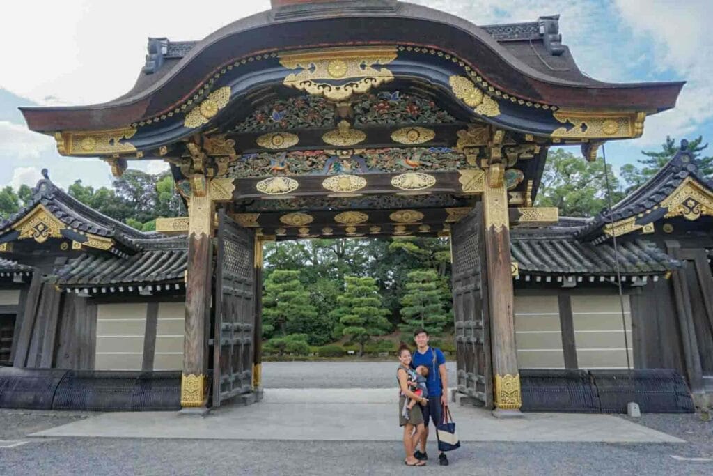 Family standing beneath an ornate wooden gate at the historic Nijo castle in Kyoto, Japan while holding their baby.