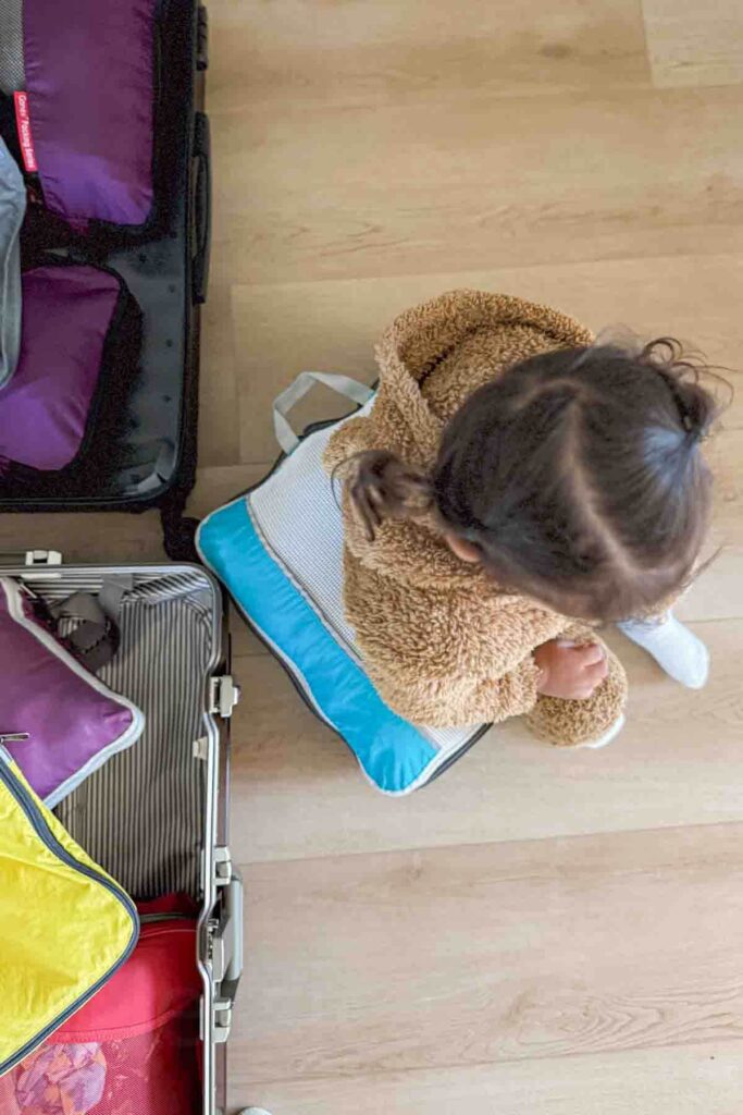 A toddler wearing a fuzzy bear style outfit sits on top of a packing cube and between two open suitcases packed with travel organization cubes.