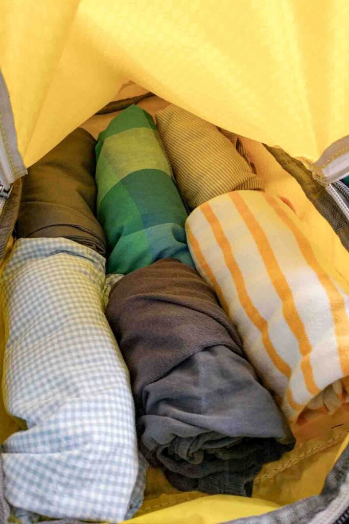 A yellow packing cube is unzipped and open wide to reveal rolled kids clothing packed tightly inside. The image demonstrates how to use stay better organized with your packing and suitcases while traveling as a family.