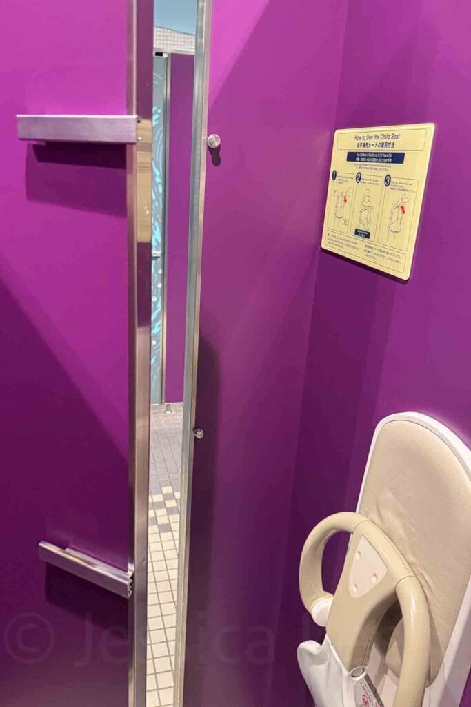 Purple public restroom stall in Japan with a fold down child seat and instruction sign showing how to use the child seat. Helpful bathroom feature when traveling to Japan with a baby.
