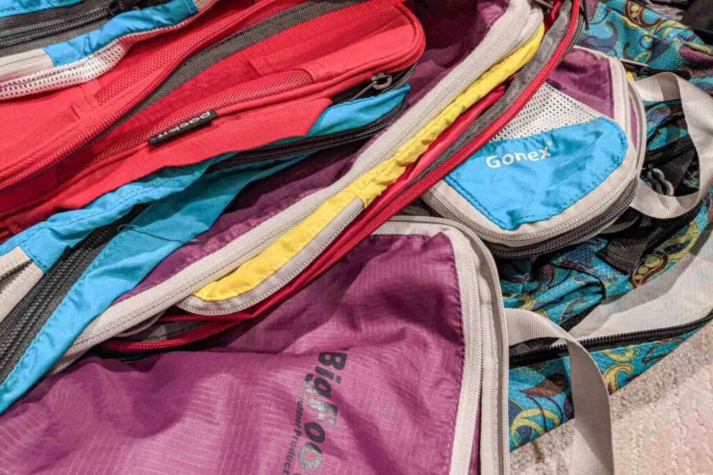 A pile of red blue and purple packing cubes is stacked together on the floor. The image reflects packing hacks for families who use color coding to organize clothing by person or category inside their suitcases.