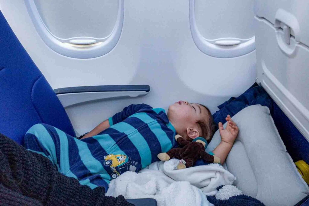Toddler in blue striped pajamas sleeping stretched across an airplane seat extended with an inflatable footrest as a makeshift bed with a pillow and blanket on a long flight, demonstrating how to get a toddler to sleep on a plane more comfortably.