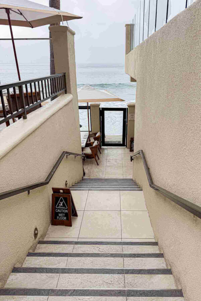 Tile staircase with a wet floor caution sign leading  down to a beachfront dining area and further down directly to the ocean. A tucked-away spot great for quiet Orange County staycations.