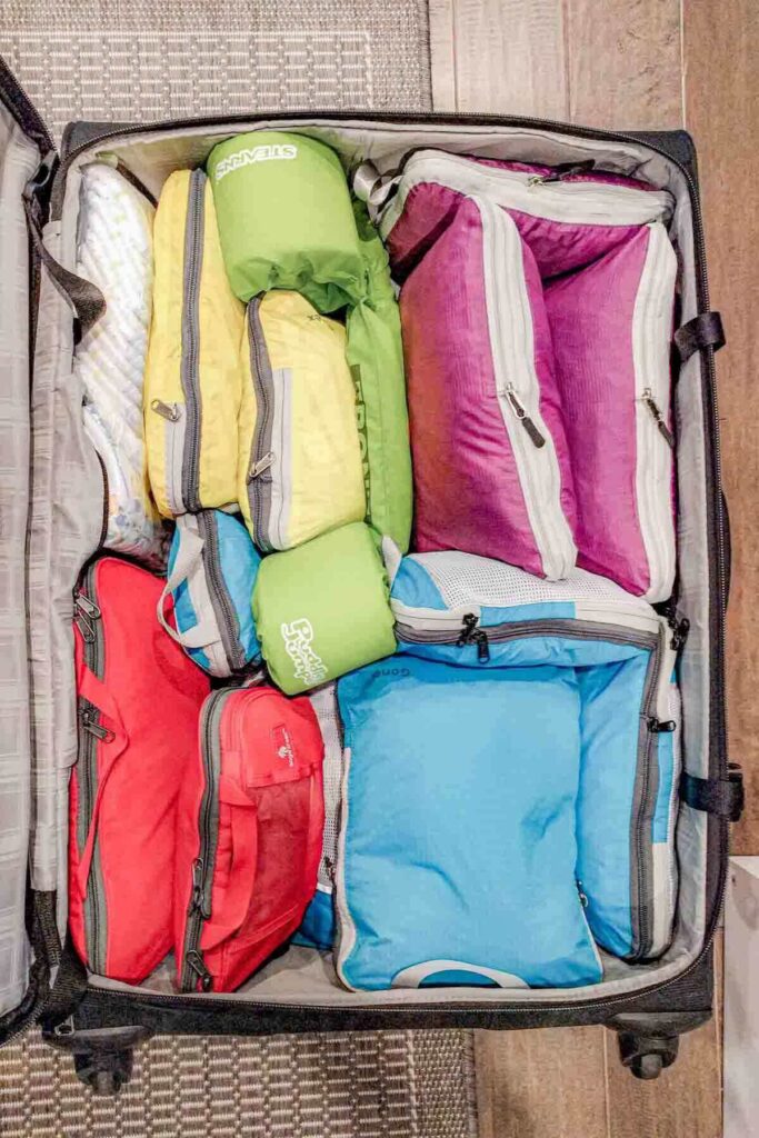 An open suitcase is completely filled with yellow, red, blue and purple packing cubes arranged edge to edge plus a green Puddle Jumper toddler life vest. This image highlights how to use packing cubes for family organization by maximizing space and keeping items separated.