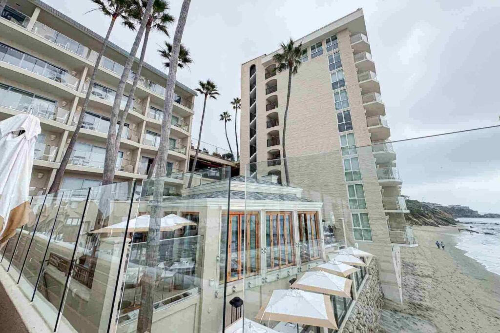 View of a beachfront hotel and glass-walled dining area with umbrellas right on top of the sand. A relaxing coastal setting for tired parents for staycations in Southern California.