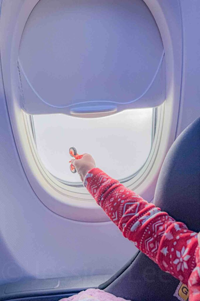 Toddler holding a small orange bike toy rolling it up an airplane window. Window seats add easy visual entertainment on a long haul flight with a toddler.