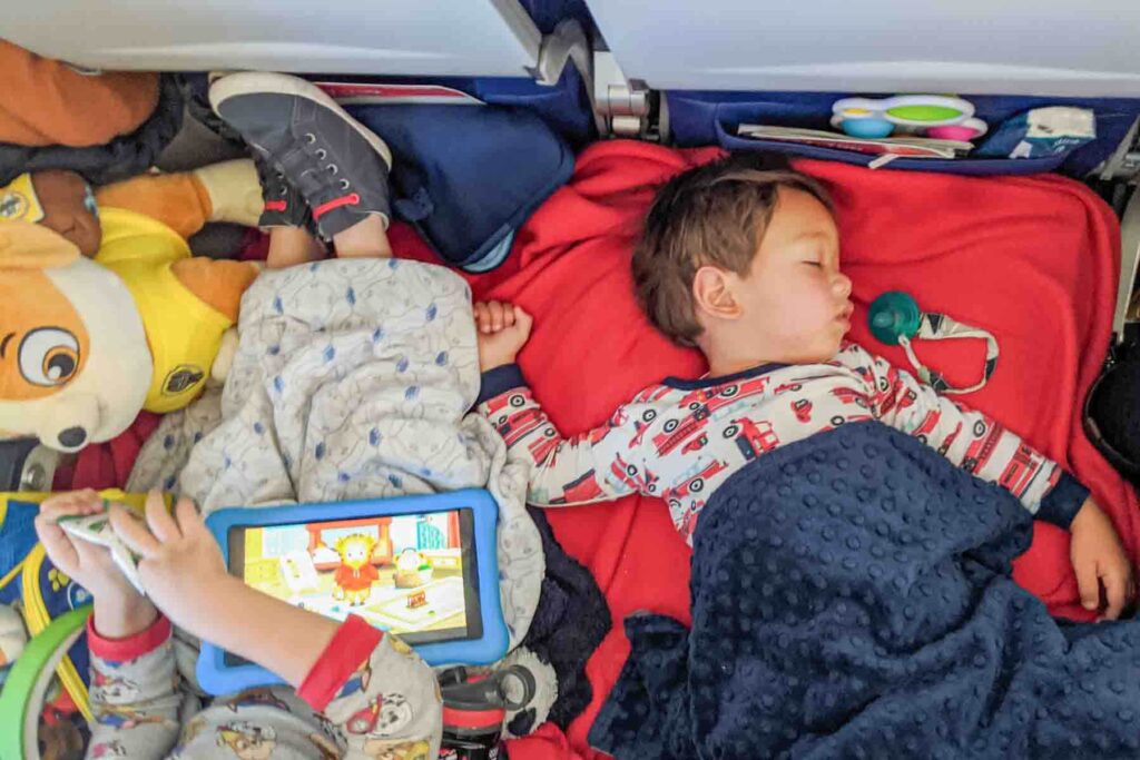 Two toddlers resting on a makeshift airplane seat bed draped with a red blanket during a long distance flight, with one asleep under a blanket and the other watching a tablet for entertainment with a Paw Patrol stuffie and backpack next to them.