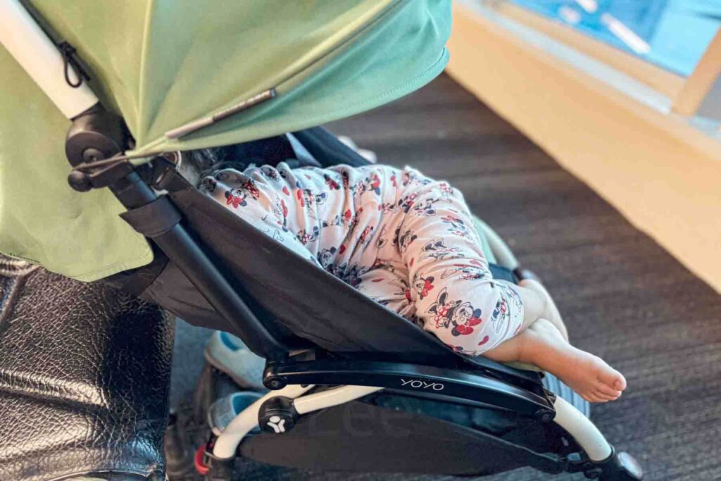 Toddler asleep in a green Babyzen Yoyo stroller at an airport during a layover wearing light pink Minnie Mouse pajamas, showing nap comfort during travel days.
