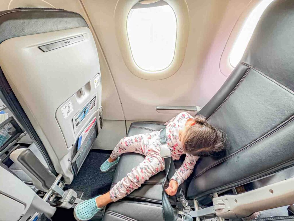 Preschooler asleep in an airplane seat with the seatbelt fastened during, showing real life how to get a toddler to sleep on a plane.
