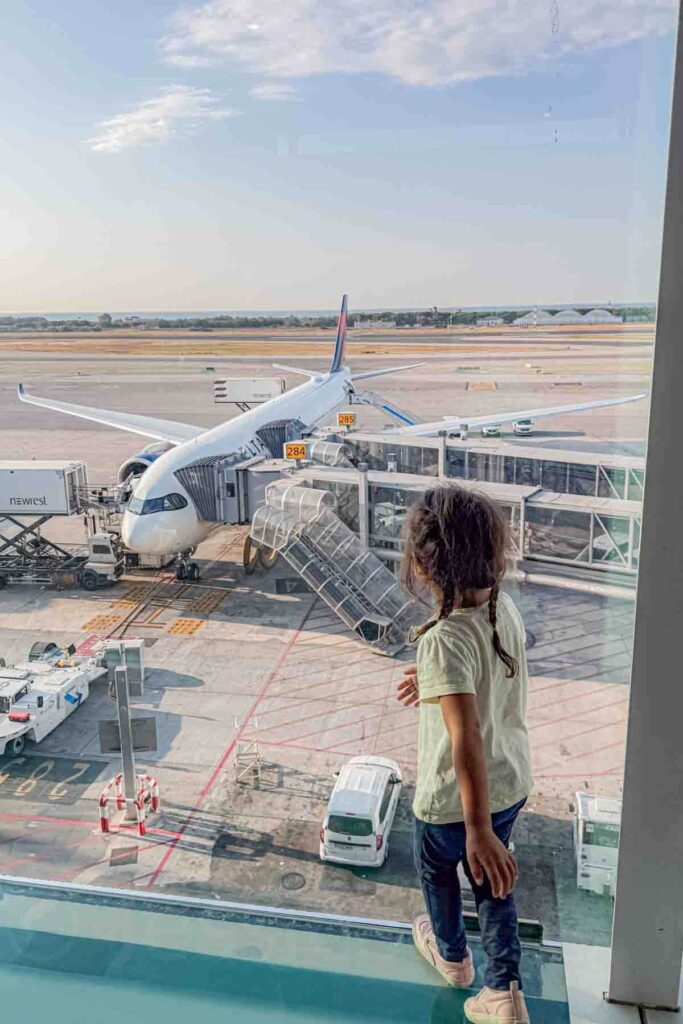 Preschooler standing at a large airport window watching an airplane at the gate before a long haul flight, building excitement before boarding.