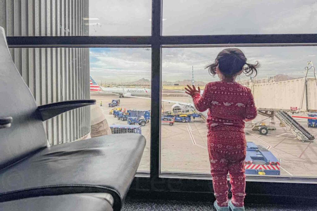 Toddler wearing red Christmas pajamas stands at an airport gate window looking out at an American Airlines plane and flight ground crew operations as she waits to board her flight.