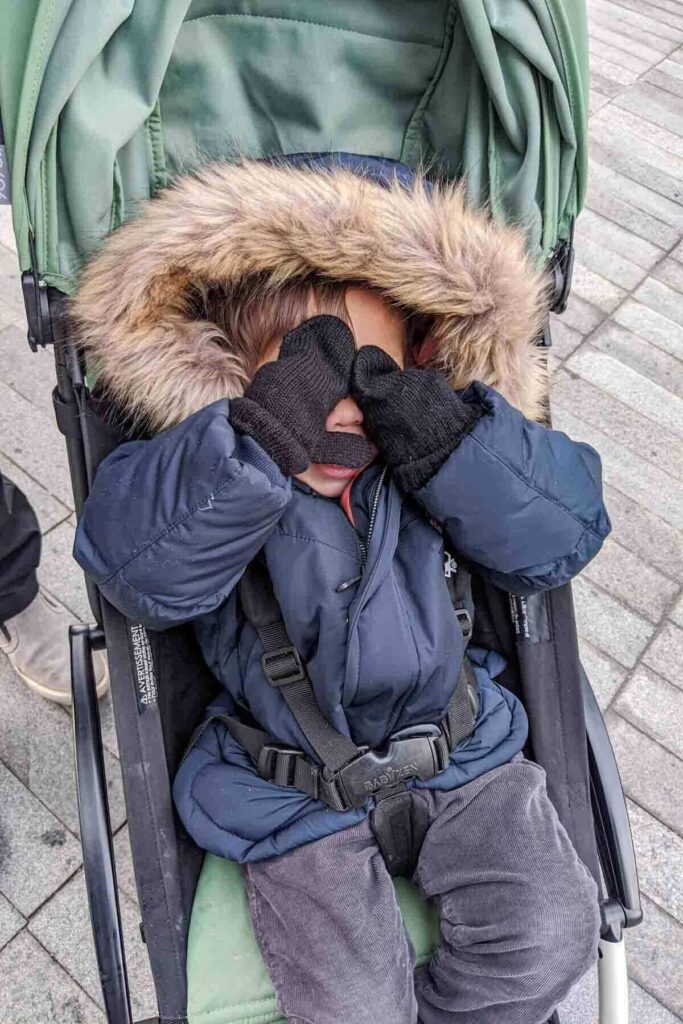 Toddler playing peekaboo while bundled in a winter coat with a giant puffy hood inside a green Babyzen Yoyo stroller, showing cold weather use while traveling.