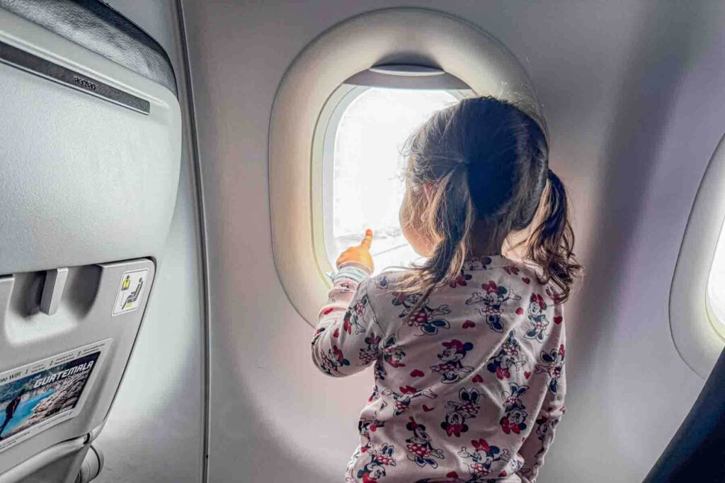 Toddler in pigtails and light pink Minnie Mouse pajamas kneeling on the seat at an airplane window and pointing outside during a flight, capturing curiosity and simple ways of entertaining a toddler on a long flight.