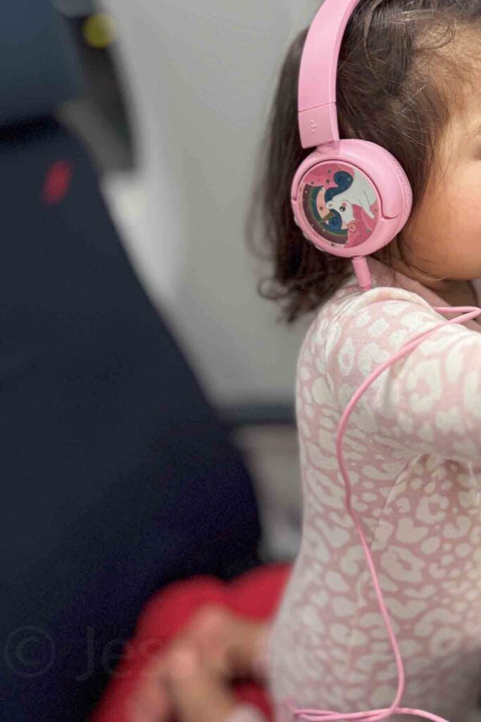 Toddler wearing pink unicorn headphones and watching a device during a long haul flight. Headphones and shows are useful for entertaining a toddler on a plane.