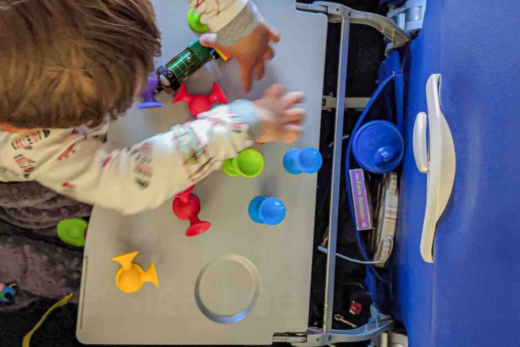 Toddler playing with Fat Brain Toyz Squigz colorful suction cups attached to an airplane tray table during a flight, showing hands on activities for toddlers on a long flight.