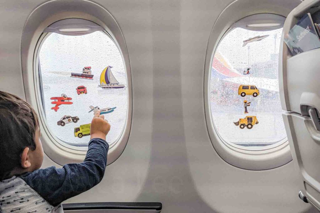 Toddler placing vehicle reusable stickers on an airplane window during a flight, showing an easy activity for entertaining a toddler on a plane.