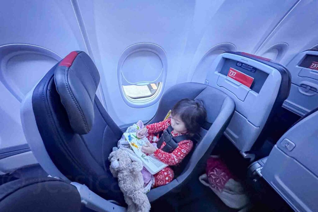 Toddler weaing red patterned Christmas pajamas sitting in a Cosco car seat on an airplane reading a board book with a stuffed animal during a long flight.