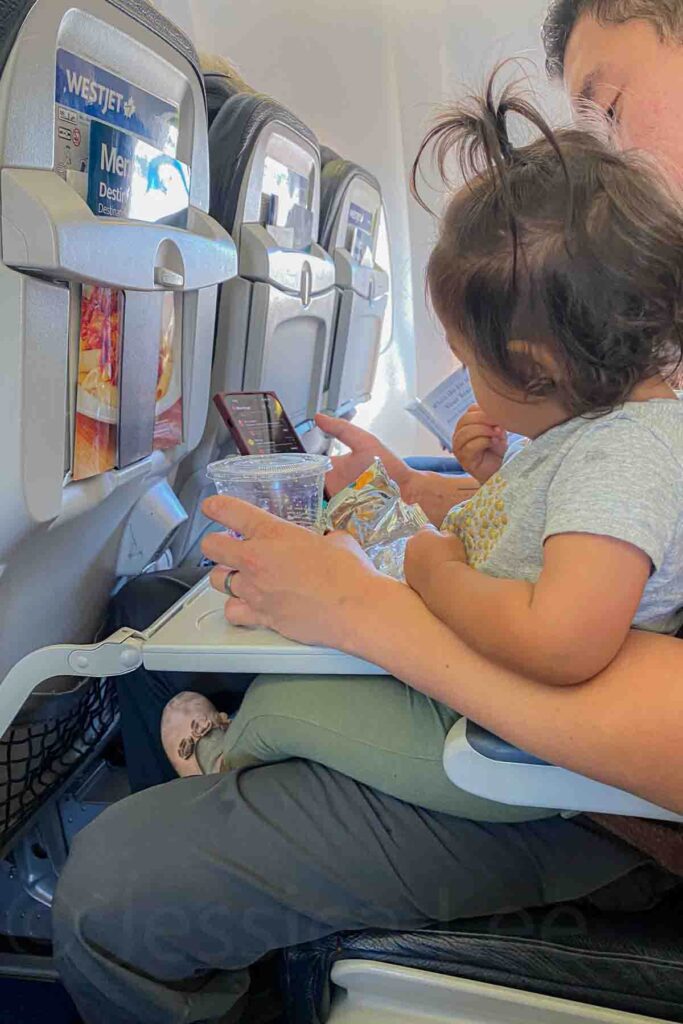 Parent holding toddler on lap on an airplane seat with snacks and a phone during a long distance flight with toddler. Snacks and screens are practical tools for entertaining a toddler on a plane.