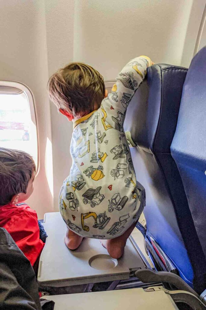 A preschooler wearing a red jacket sits in an airplane seat next to the window with his toddler brother next to him wearing white and yellow construction truck pajamas squatting on an airplane tray table looking out the window during a flight with a toddler, showing toddlers need to move.