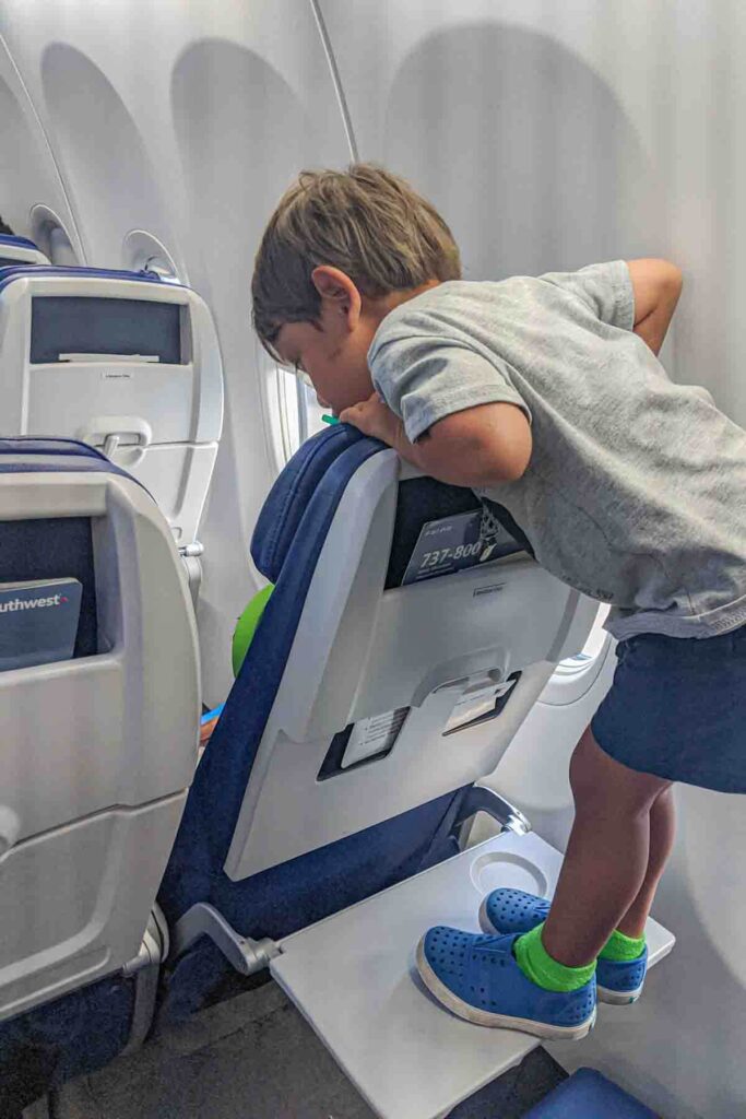Toddler standing carefully on an airplane tray table and looking over the seat in front of him during a flight. 