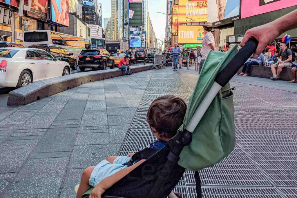 Toddler riding in a green Yoyo travel stroller watching the bright lights of Times Square in New York City, showing crowd navigation in a busy urban setting.