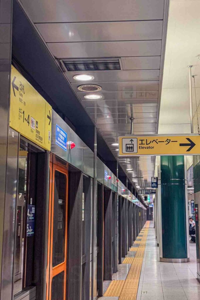 Yellow elevator sign with Japanese and English text hanging above a Tokyo subway platform. Useful accessibility feature when exploring Tokyo with a baby.