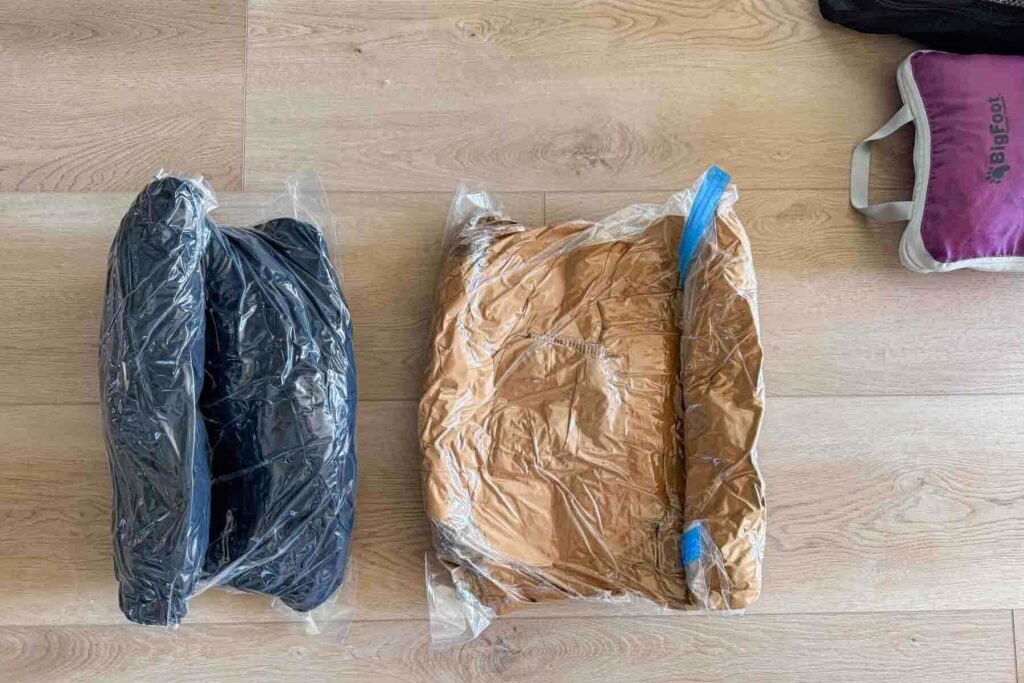 Winter clothing is sealed inside clear vacuum compression bags laid flat on a wooden floor. The image highlights packing hacks for families who need to save space when traveling with bulky seasonal clothing. A couple packing cubes are visible in the top right corner of the image.